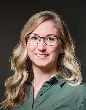Professional headshot of a smiling blonde woman with glasses wearing a green shirt against a dark background.