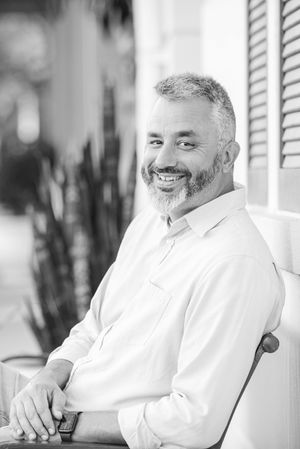 Black and white portrait of a smiling middle-aged man with a beard sitting outdoors in a chair.