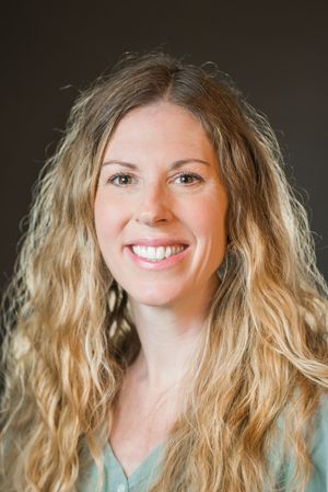 Professional headshot of someone with wavy blonde hair and bright smile wearing a teal top against dark background.