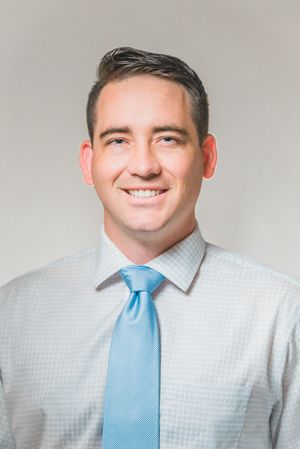 Professional headshot of a smiling young man in a light blue tie and checkered dress shirt against a gray background.