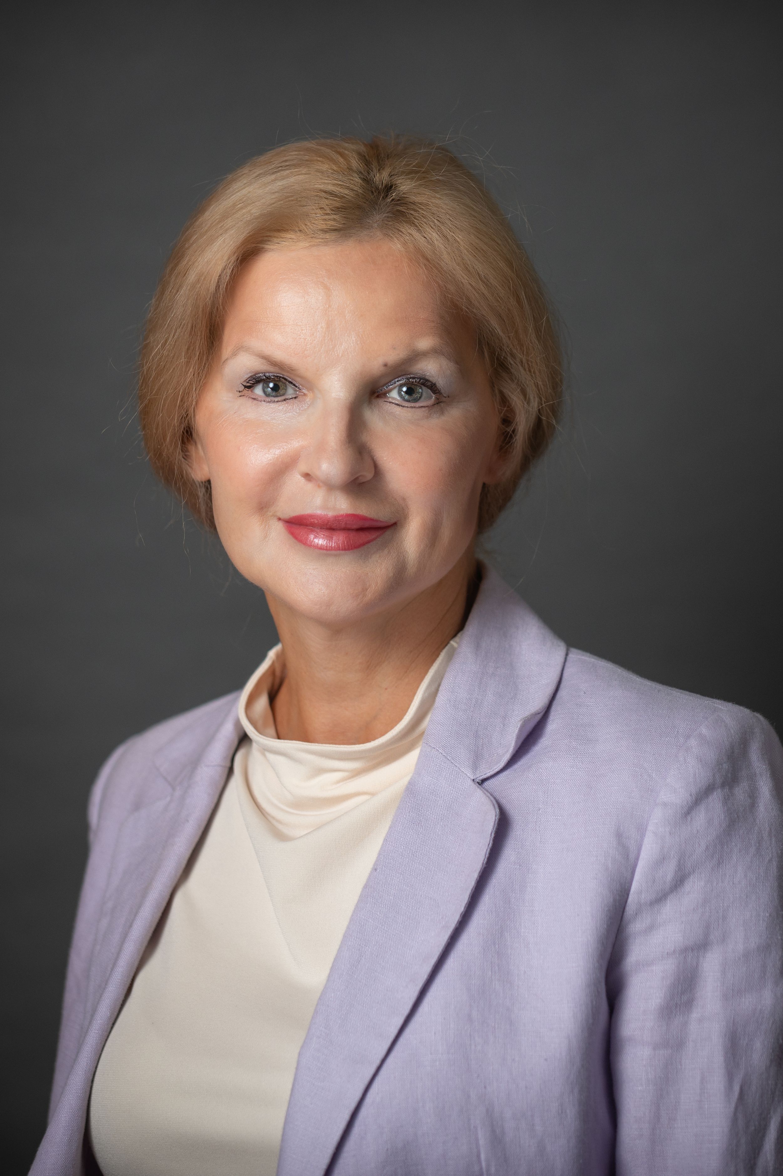 Professional headshot of a mature blonde woman in a lavender blazer smiling confidently against a gray background.