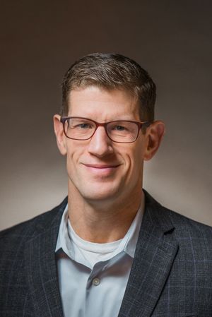 Professional headshot of person in grey suit and glasses against neutral background.