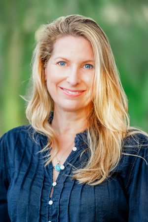 Smiling blonde woman with blue eyes wearing a navy blouse and turquoise necklace outdoors with green background.