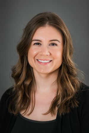 Professional headshot of a smiling woman with long brown hair wearing a black top against a gray background.