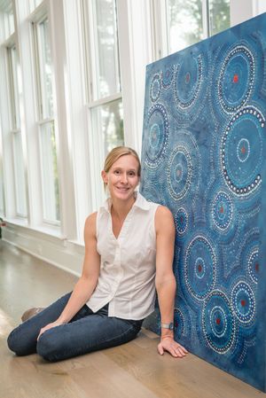 Smiling blonde woman sitting cross-legged on hardwood floor beside a large blue Aboriginal dot art painting.