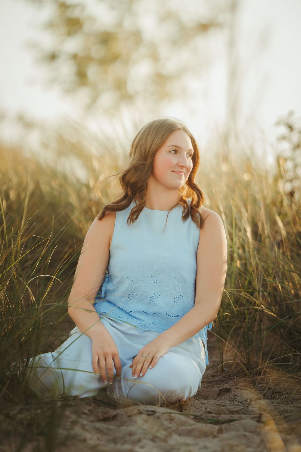 Teen girl in blue outfit sitting on sandy beach ground surrounded by tall grass.