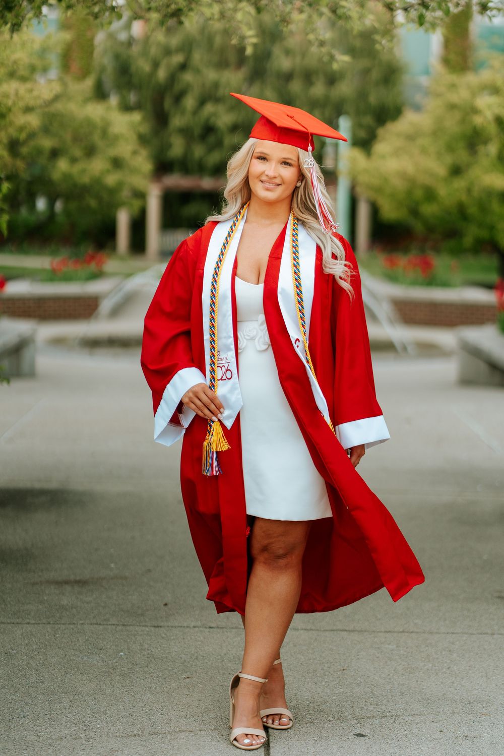 Smiling graduate in red cap and gown with gold honor cords poses outdoors in white dress and heels.