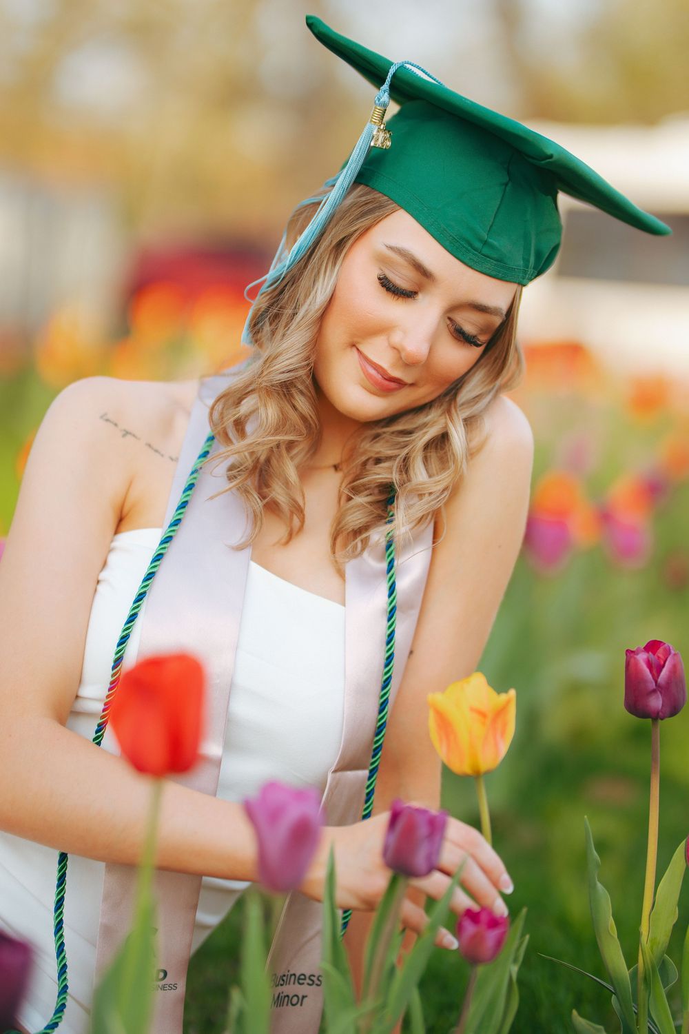 Smiling graduate in green cap and white dress admiring colorful tulips in a vibrant flower field.
