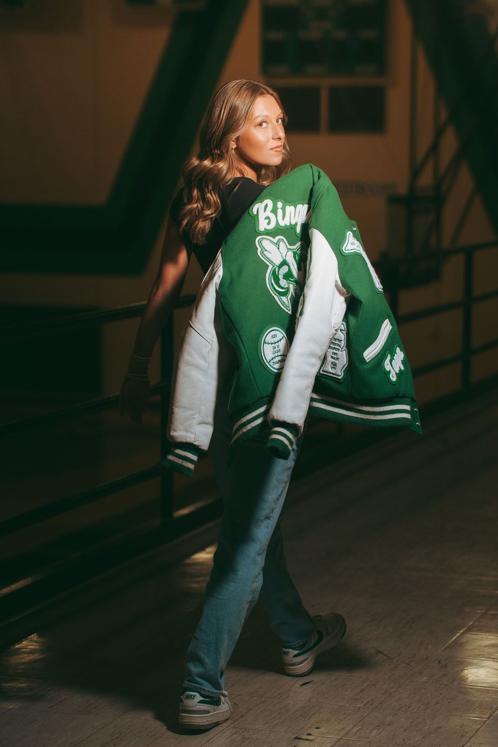 Young woman posing in green varsity jacket and jeans in dimly lit indoor setting.