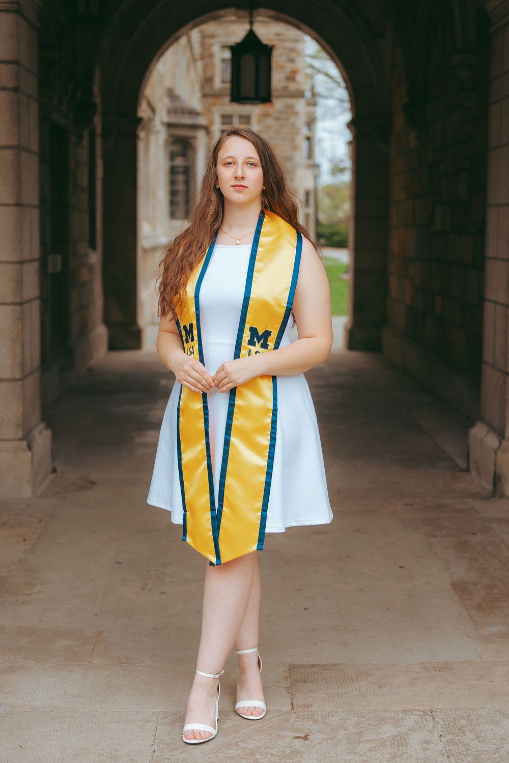 Young woman in white dress wearing University of Michigan gold graduation stole poses under stone archway.