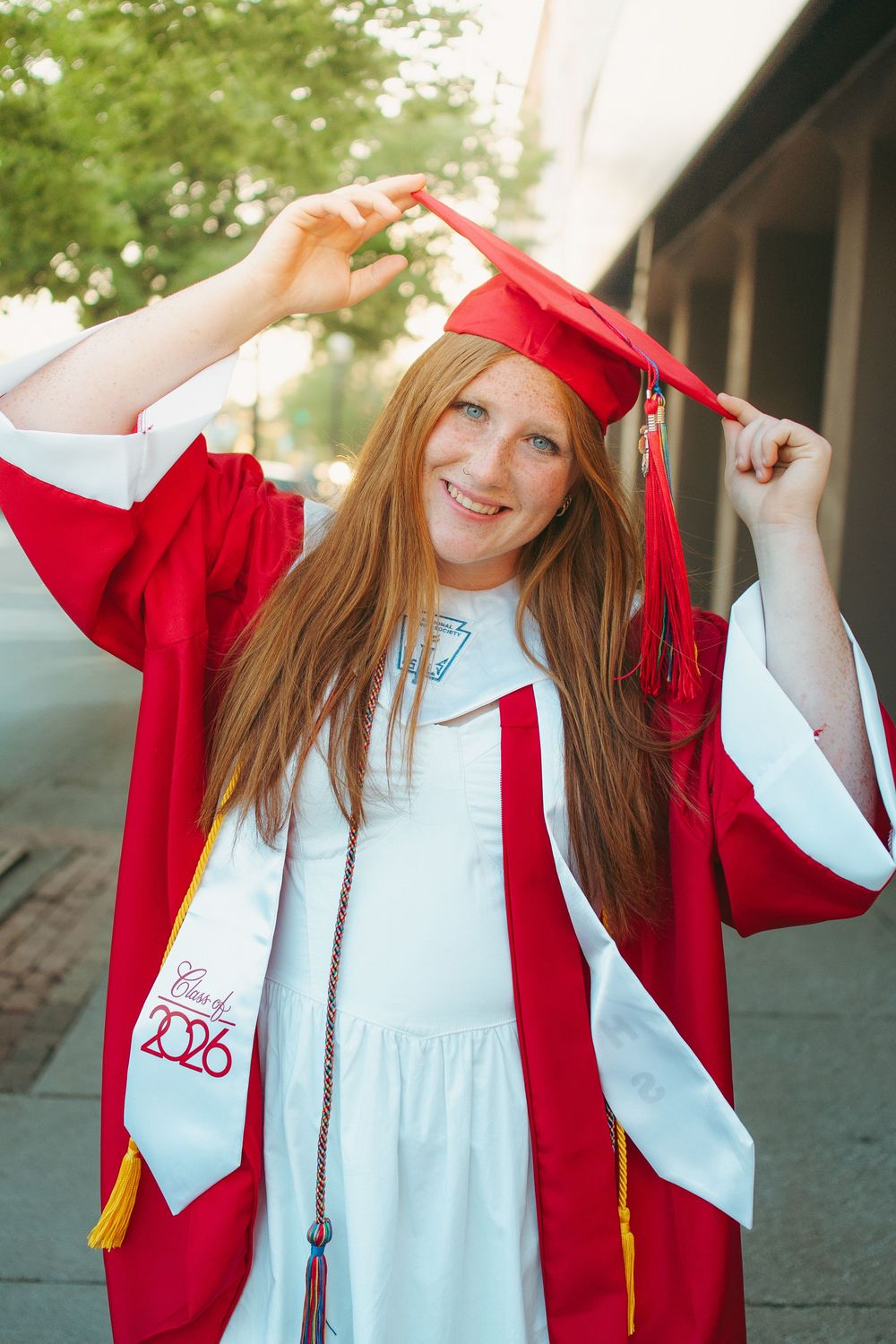 Smiling redhead graduate in red cap and gown, Class of 2026, posing outdoors on a sunny day.