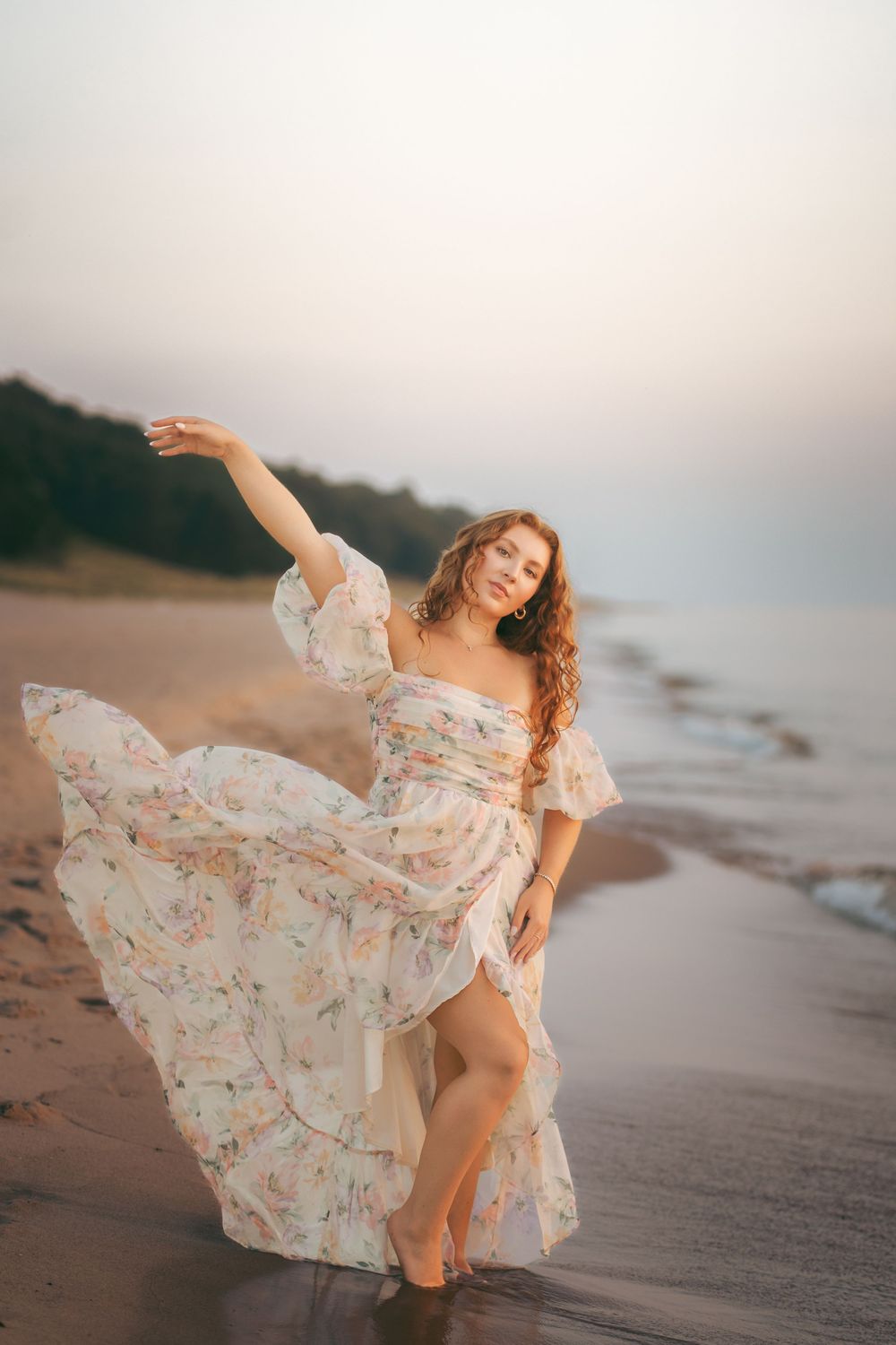 Woman in a flowing floral dress dancing gracefully on a sandy beach at sunset by the water.