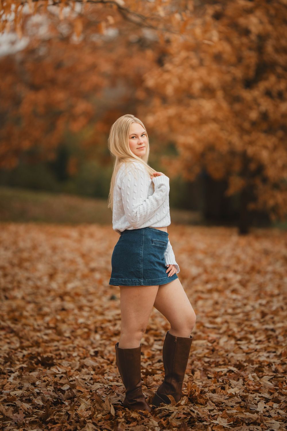 Blonde teen girl in white sweater and denim mini skirt standing in fall leaves wearing brown boots.