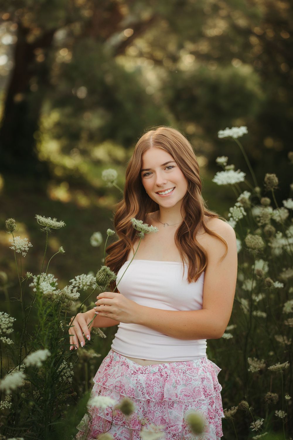 Smiling red-haired teenage girl in a white top and pink skirt standing among white wildflowers in a forest.