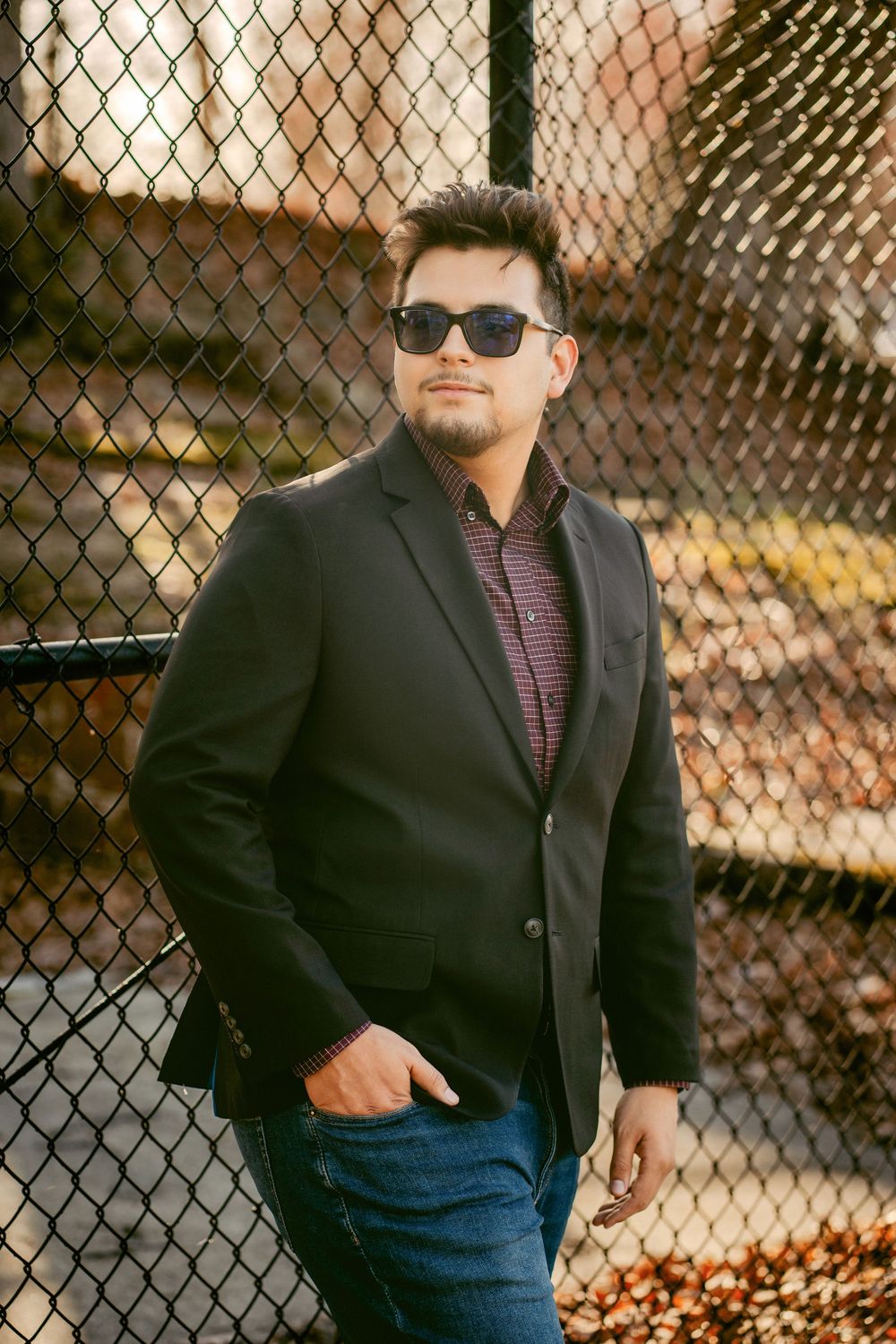 Man in black blazer and sunglasses posing confidently by a chain-link fence in autumn setting.