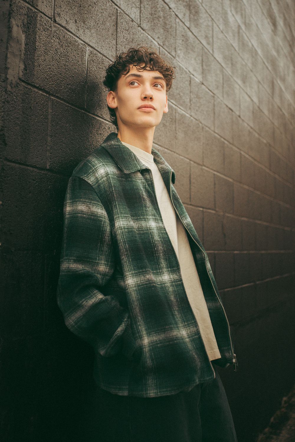 Young man in green plaid jacket leaning against dark brick wall, looking up thoughtfully in natural light.