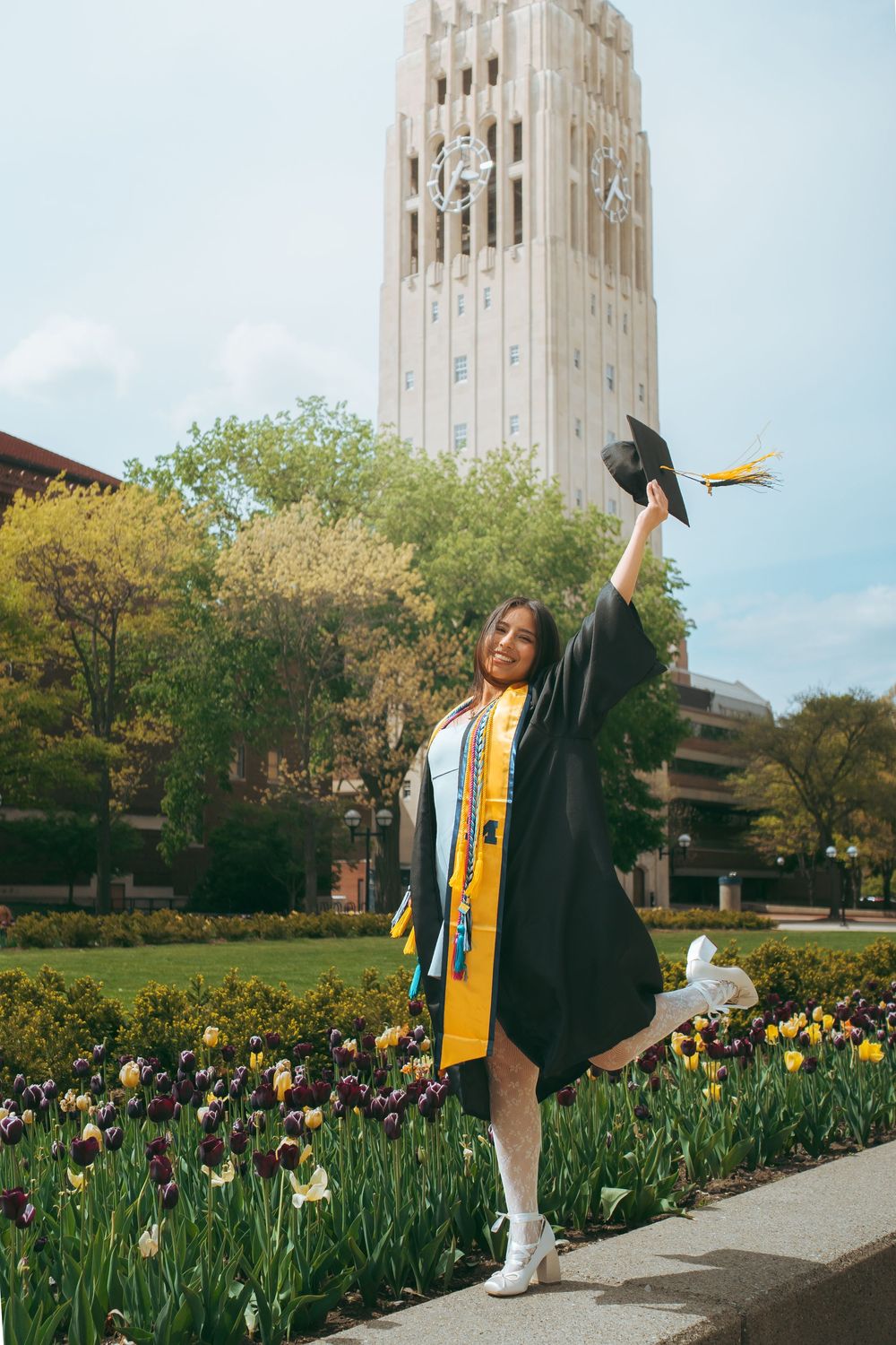 A smiling graduate in black cap and gown with gold sash raises her cap near a university bell tower and tulip garden.