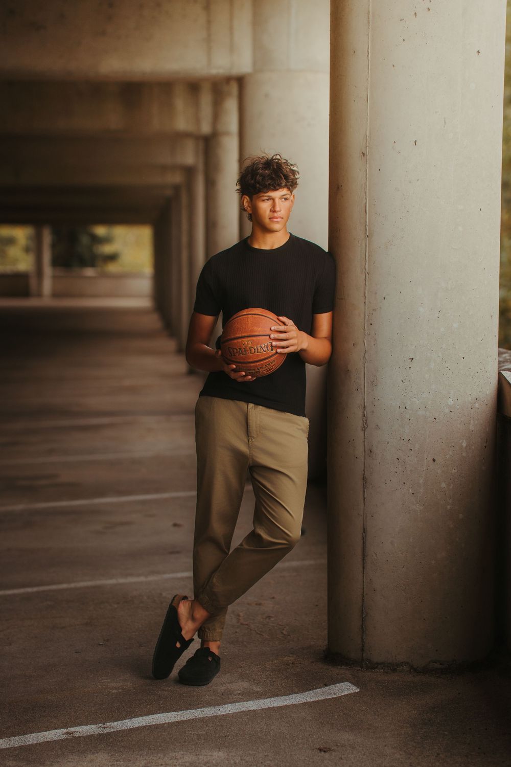 Teen boy in black tee and khakis holding basketball leaning against concrete column in parking garage.