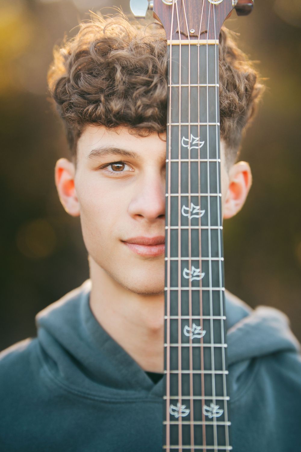 Young curly-haired teen holding a guitar neck vertically in front of his face outdoors at golden hour.