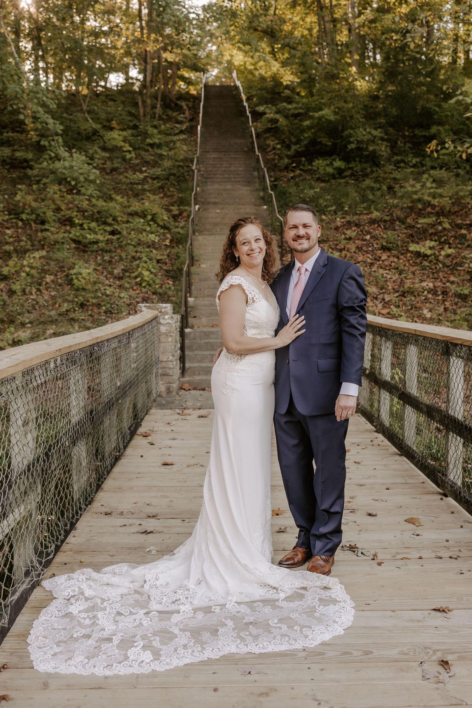 A couple in wedding attire poses on a wooden bridge surrounded by autumn foliage during their outdoor ceremony.