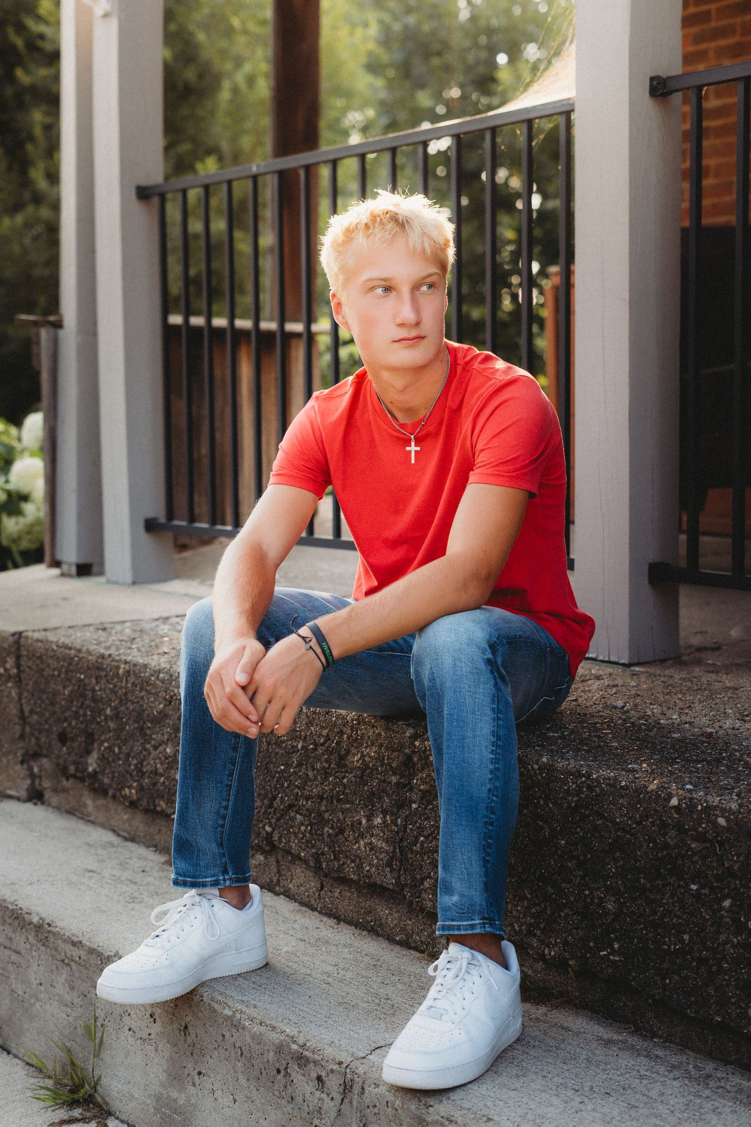 Blonde teenage boy in red t-shirt, jeans, and white sneakers sits on stone steps for senior portrait.