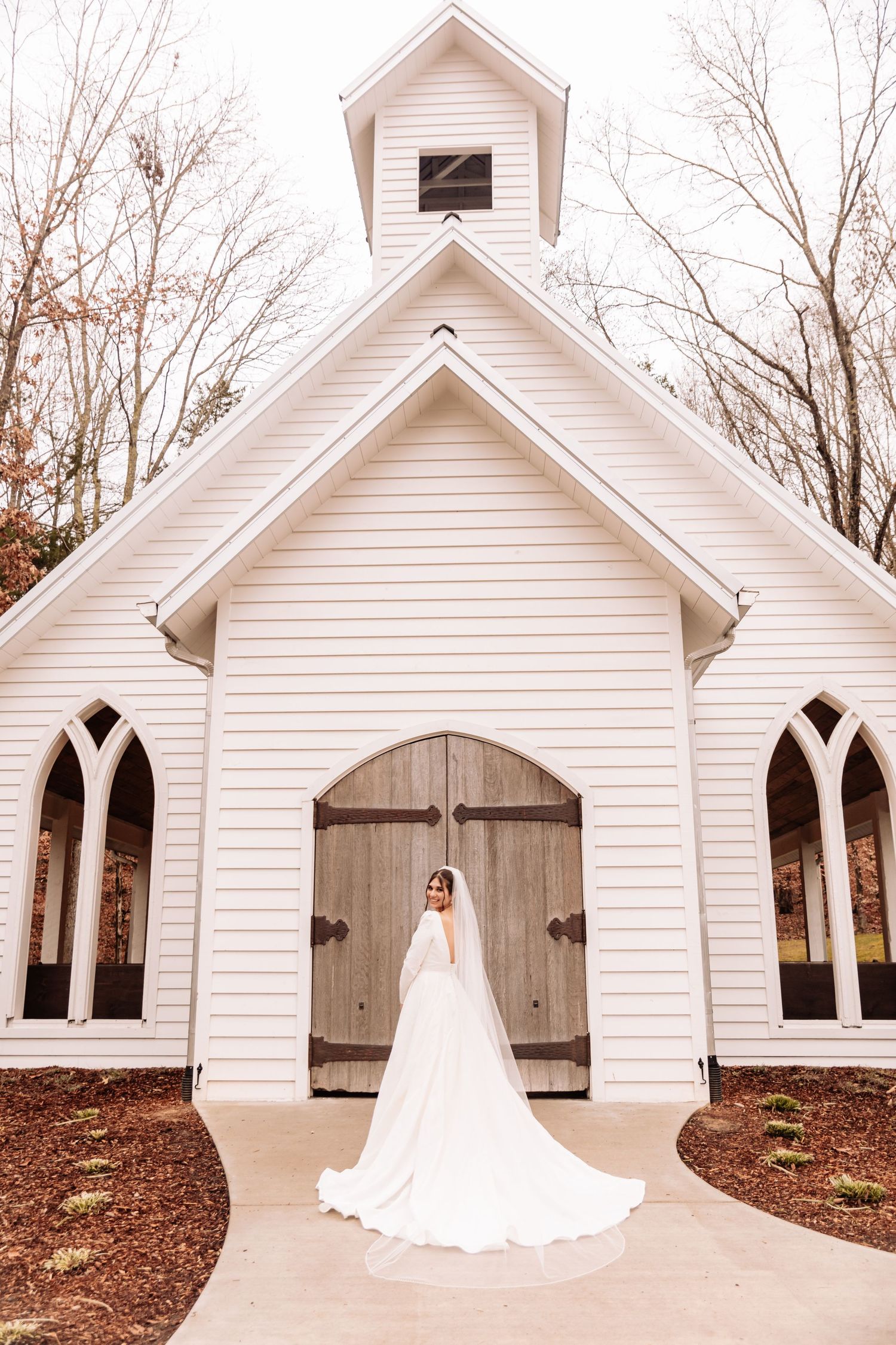 A white wooden chapel with Gothic windows and rustic wooden doors stands among bare trees on an autumn day.