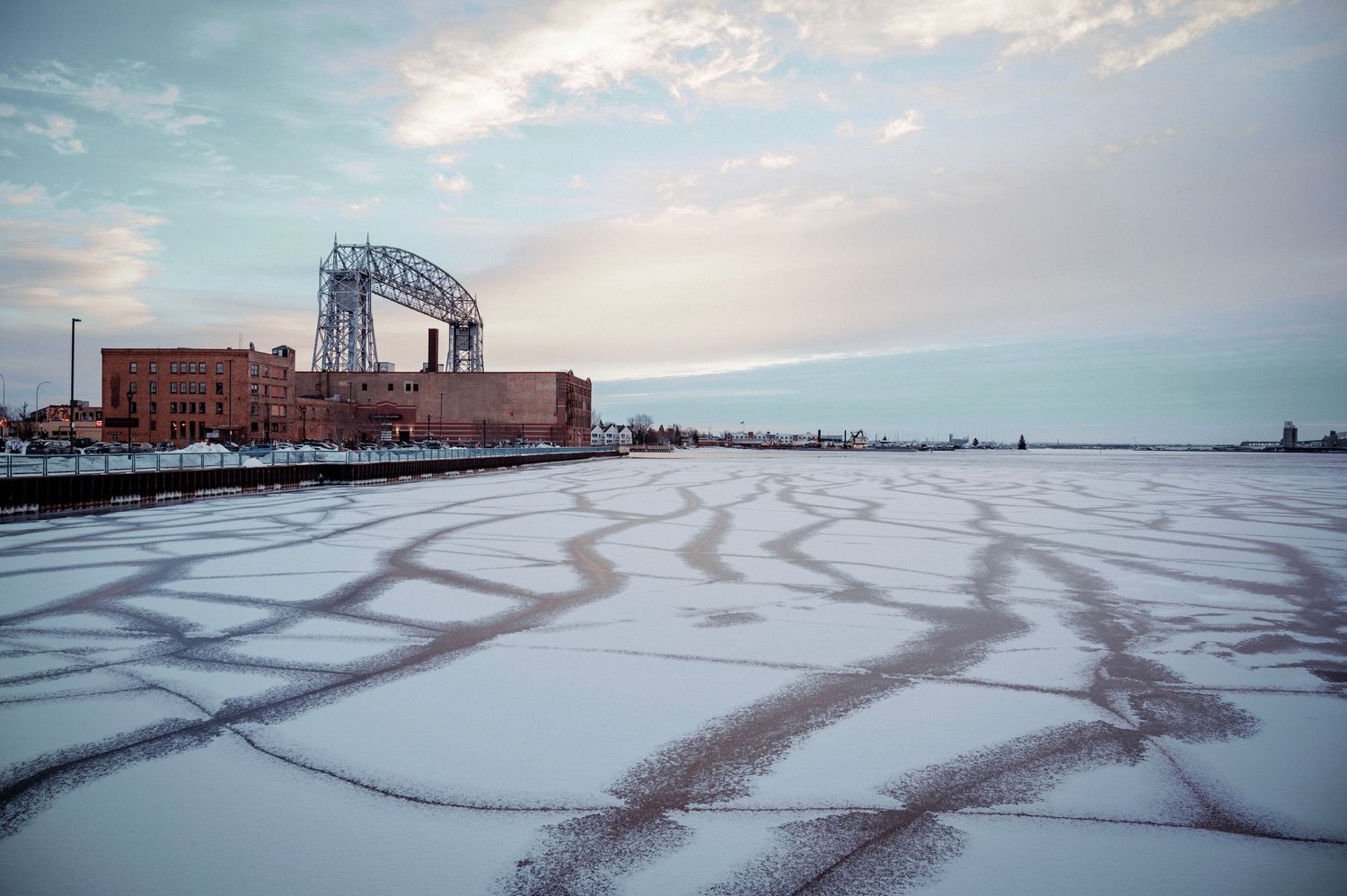 Winter panorama of Lake Superior shoreline with tire tracks in snow and industrial buildings.
