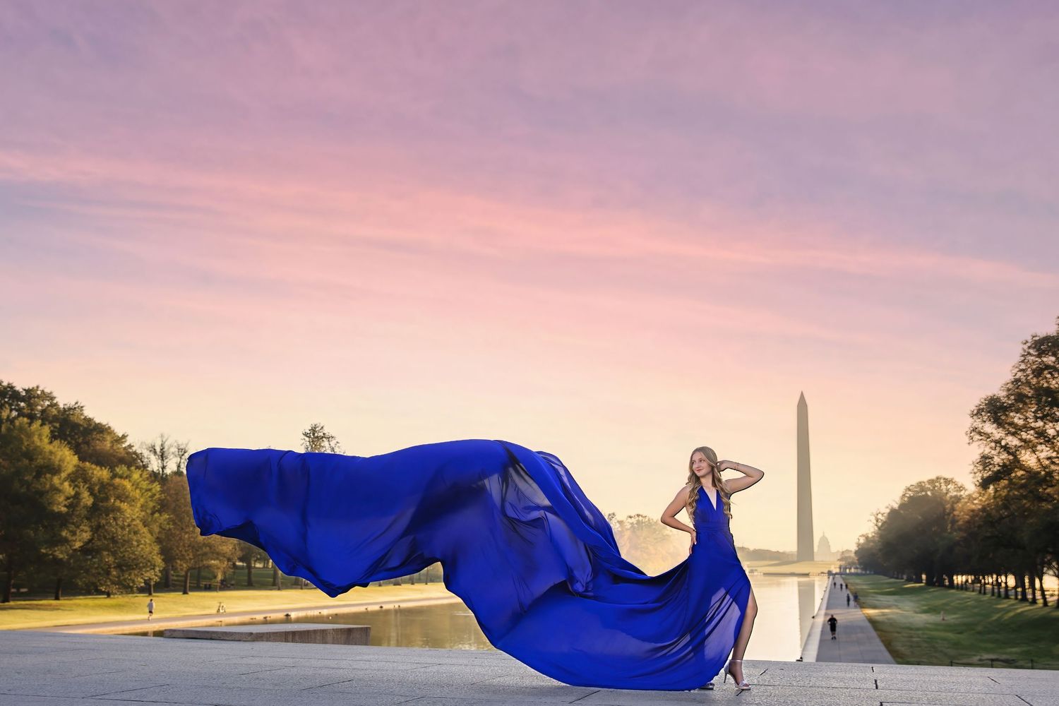 A flowing blue fabric billows gracefully against a sunset sky with monuments and trees silhouetted in the background.