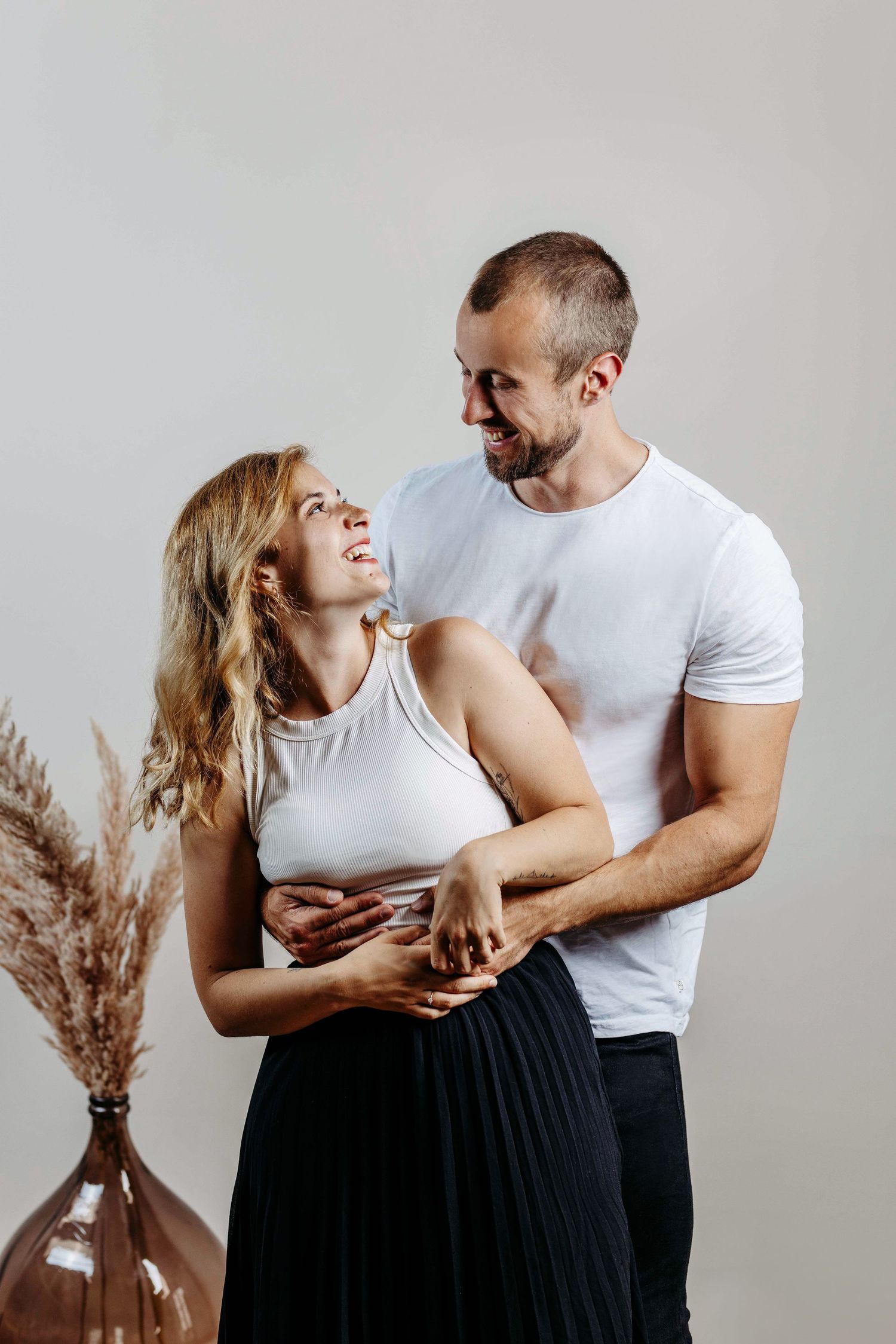 A romantic couple in white tops and black bottoms share tender moments in a minimalist studio setting with pampas grass decor.