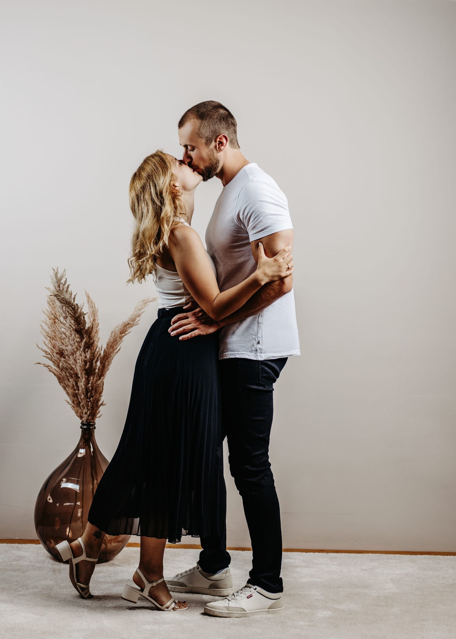 A romantic couple in casual attire share intimate moments in a minimalist studio setting with dried pampas grass decor.
