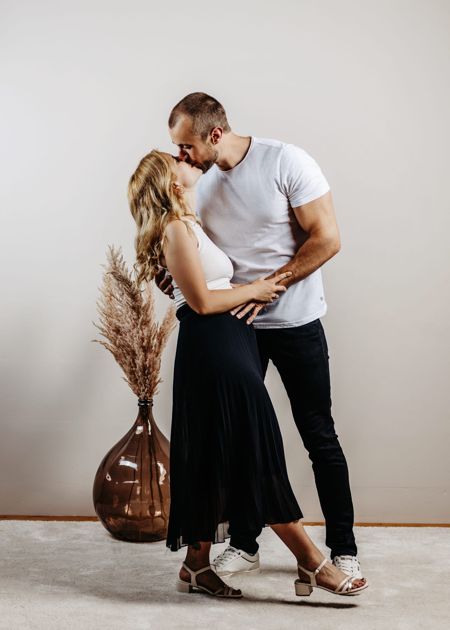 A romantic couple in casual attire poses together in a minimalist studio setting with a brown vase and pampas grass decor.