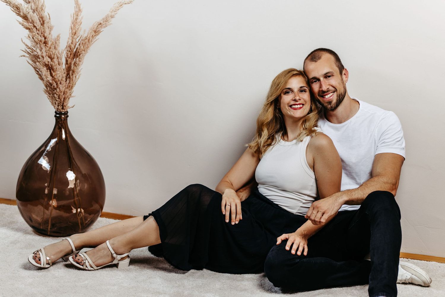 A couple sits on the floor beside a brown vase with pampas grass, wearing casual white and black outfits.