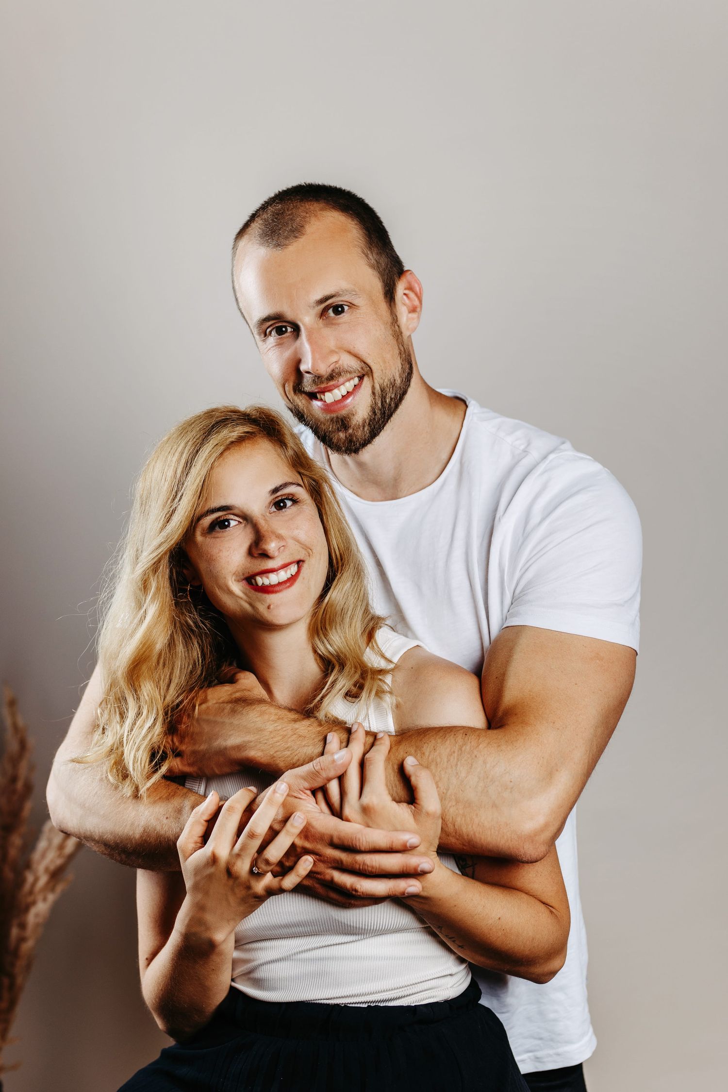 A couple embraces in a casual portrait against a neutral background, wearing white and sharing warm smiles.