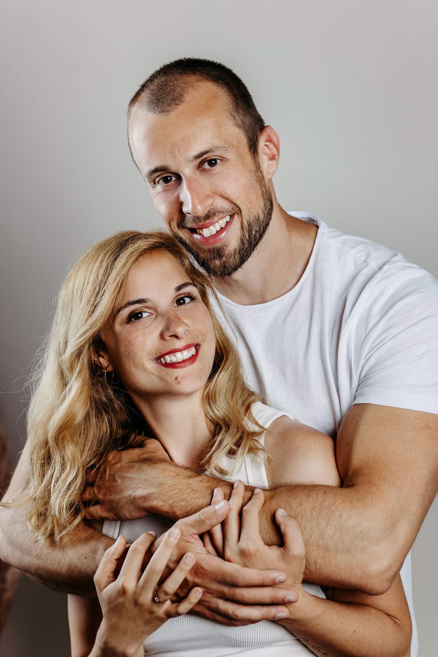 A couple in white shirts embrace and smile warmly against a neutral background in an intimate portrait.