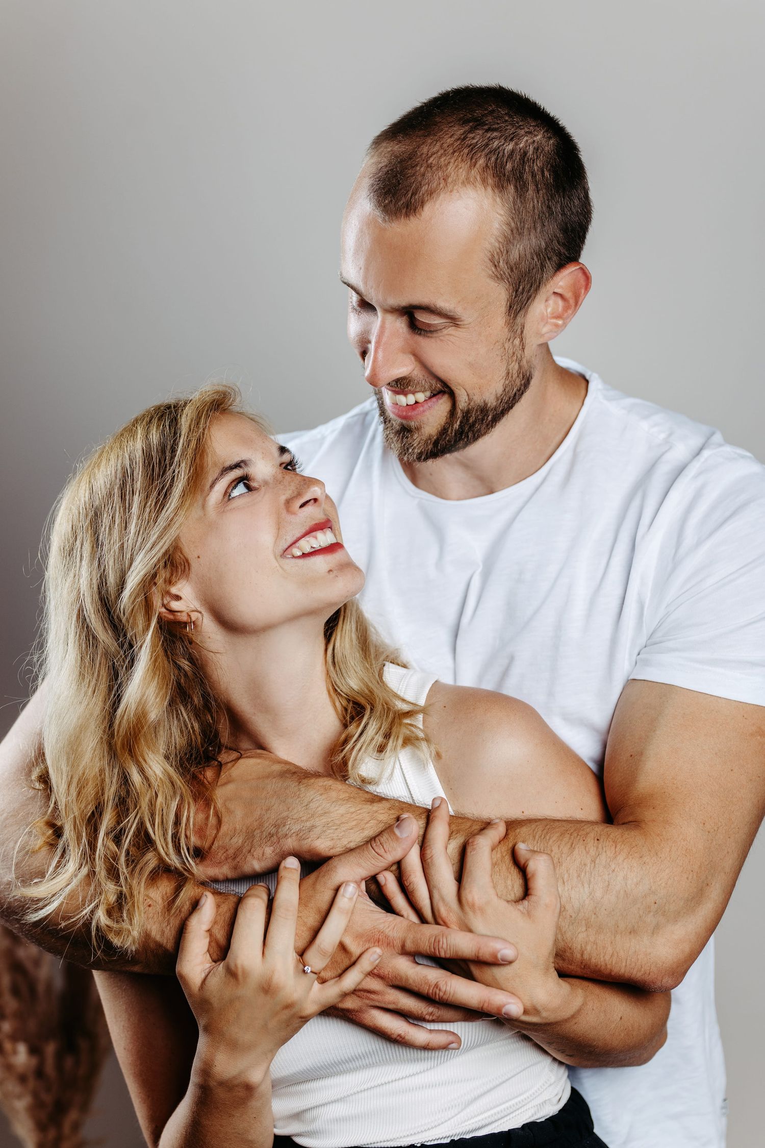 A couple in white shirts sharing a joyful embrace and laughing together against a gray background.
