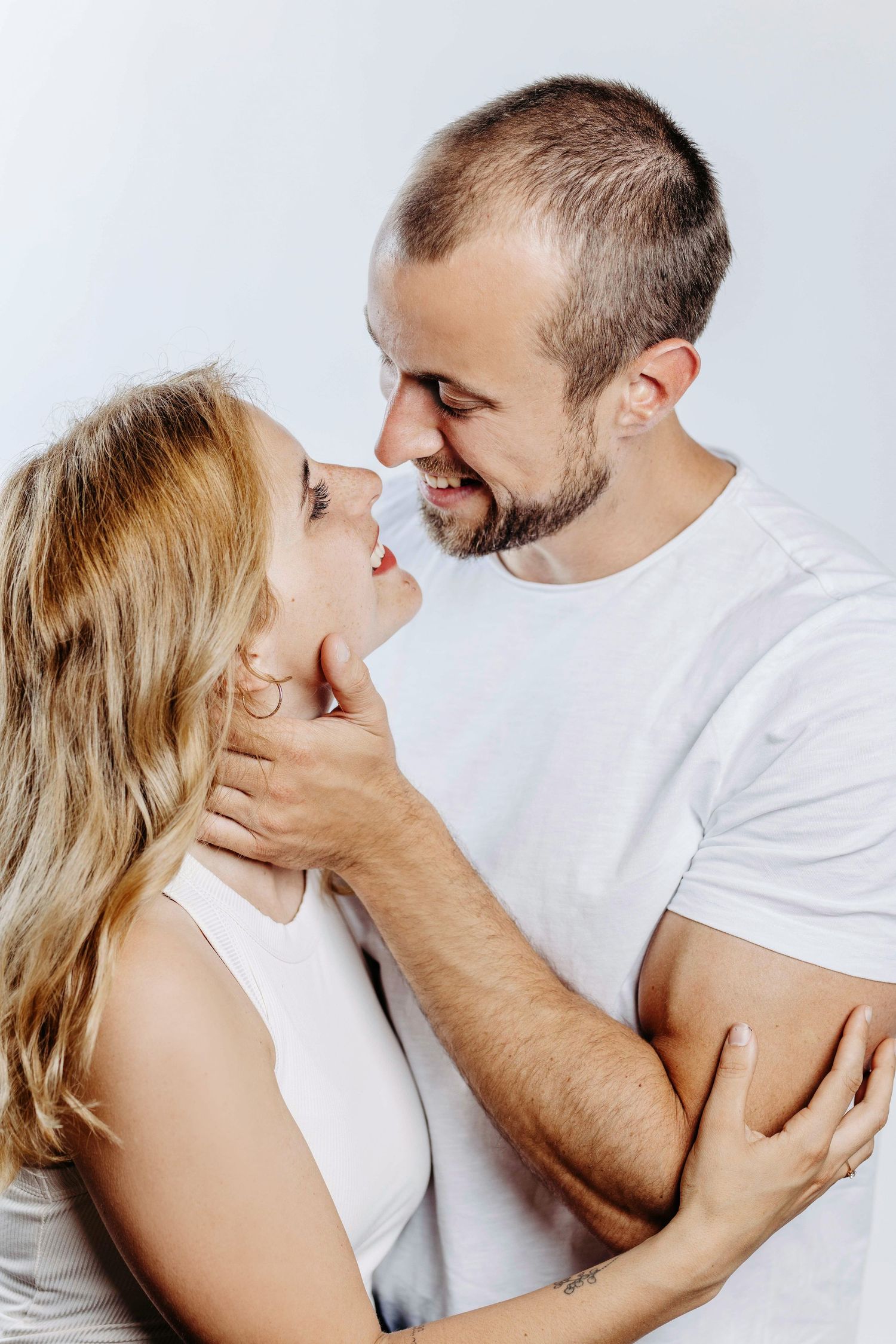 A romantic couple in white clothing sharing an intimate moment while embracing against a light background.