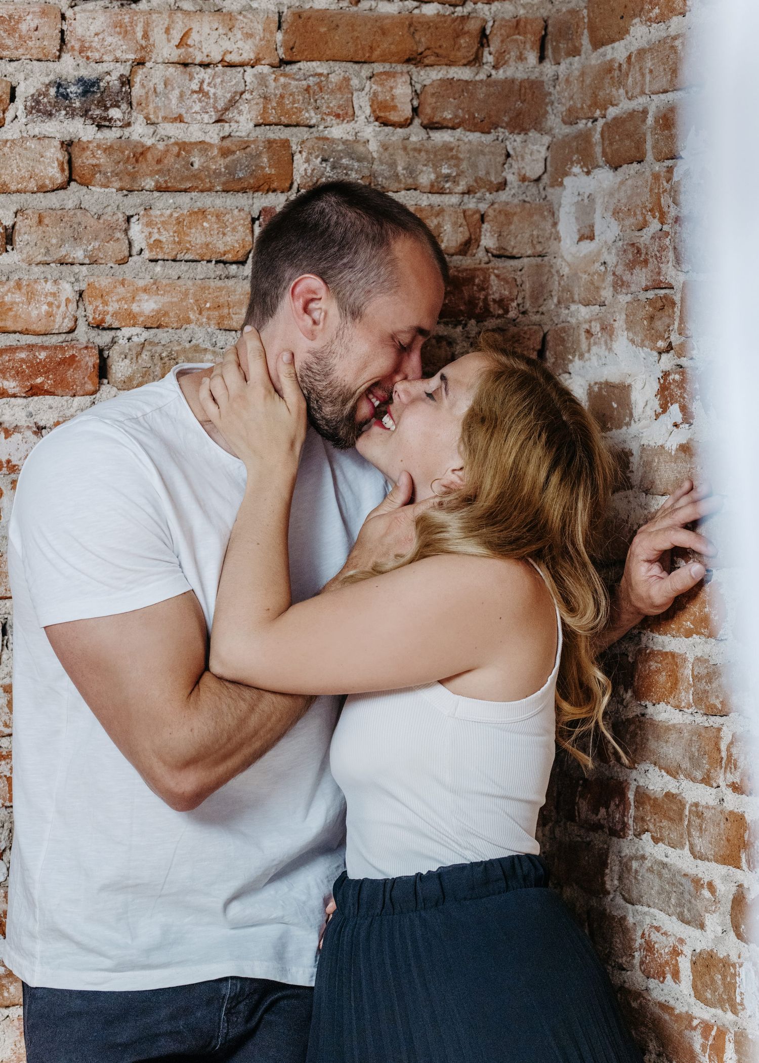 A romantic couple in white and navy outfits share intimate moments against a rustic exposed brick wall.