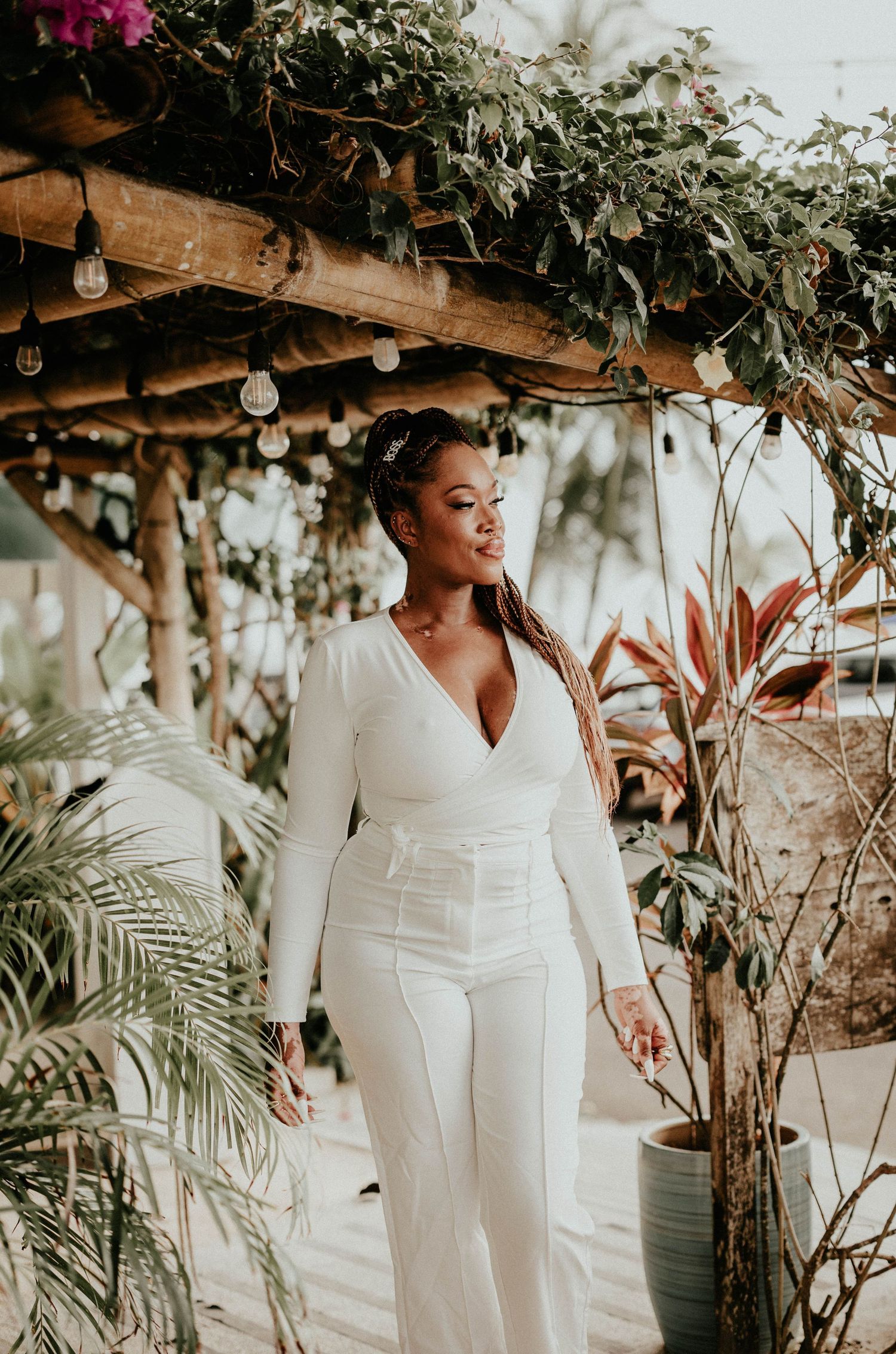 A person in an elegant white wedding jumpsuit poses under rustic wooden beams decorated with greenery and string lights.