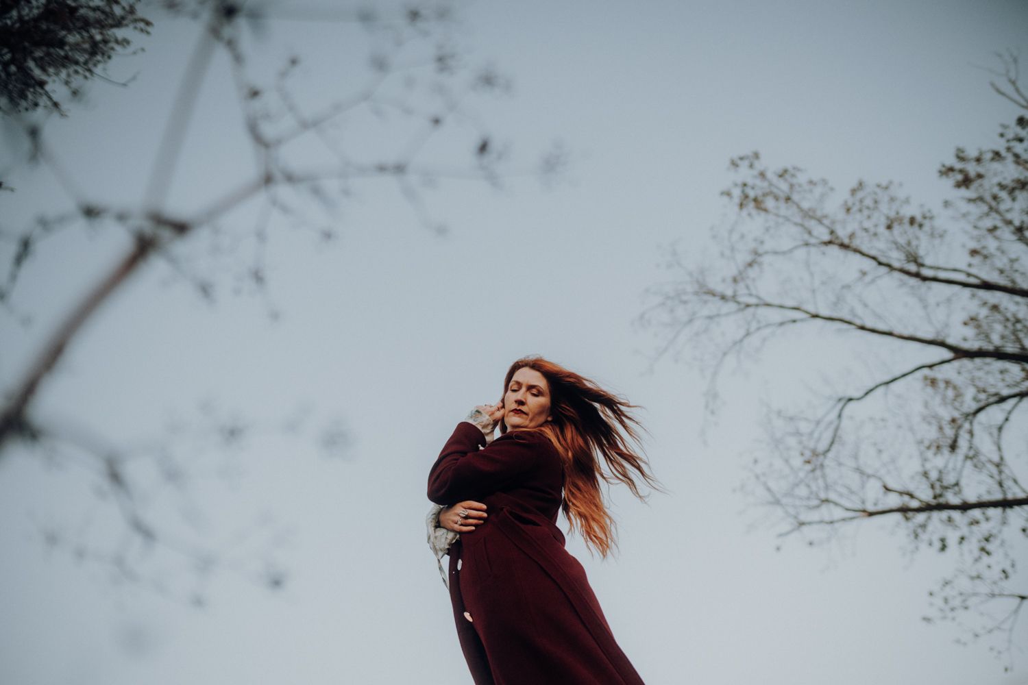 Portraitshooting mit Hey Elli Fotoreportagen an der Elbe. Es ist windig und ihr rotes Haar fliegt im Wind. Der Himmel ist bewölkt.