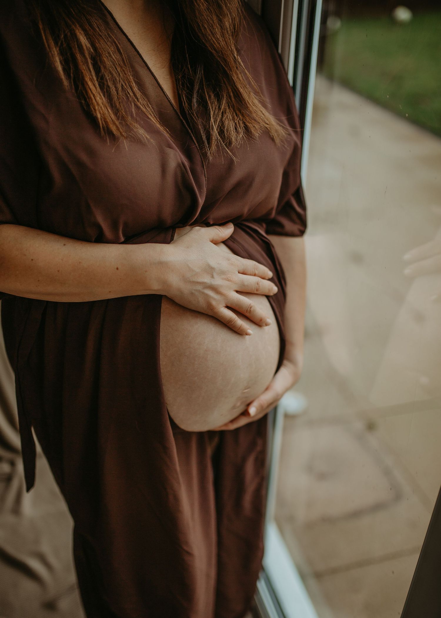 Close-up view of pregnant woman in brown dress cradling her baby bump near a window.