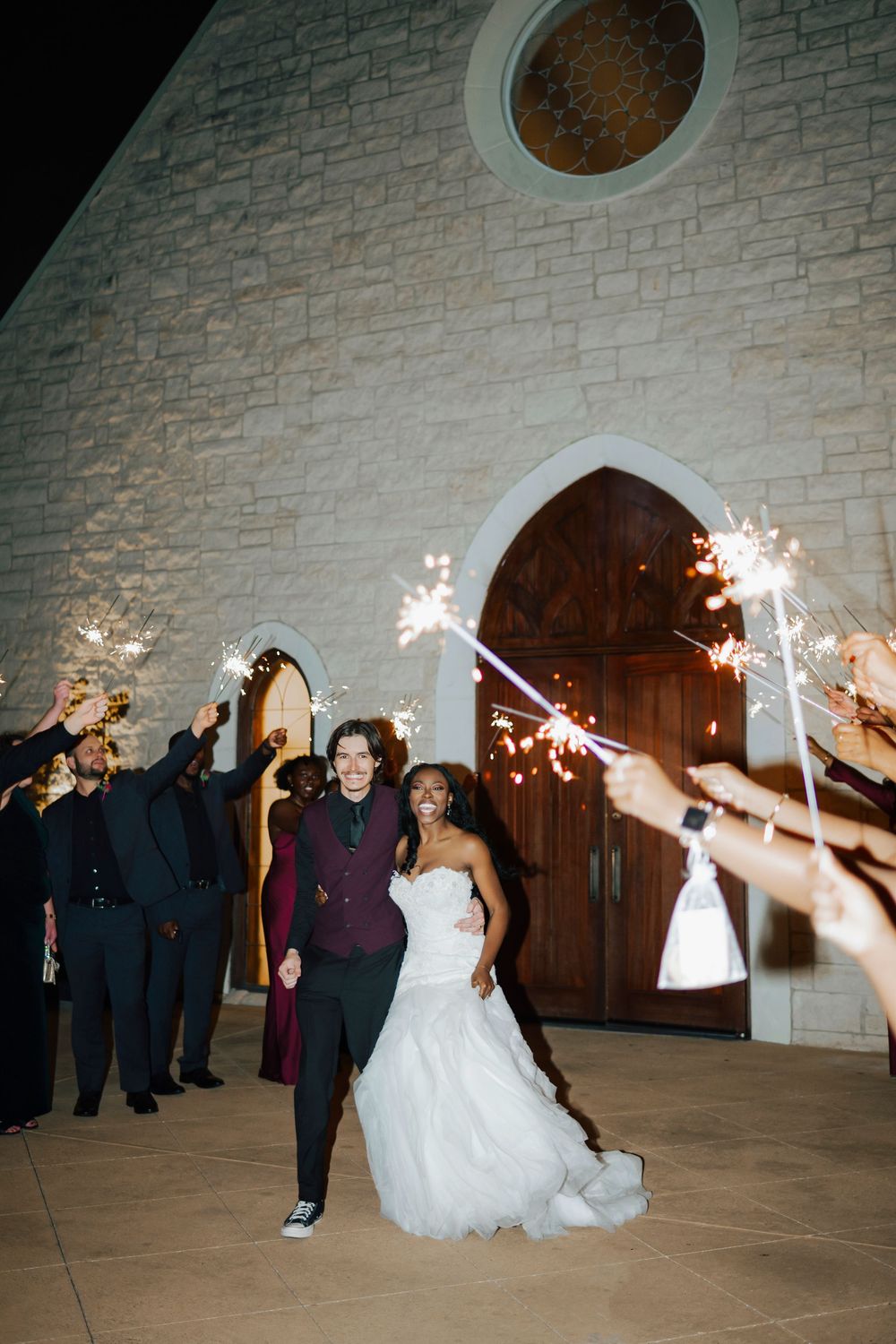 Wedding party members hold sparklers in a curved formation creating a magical tunnel effect at an evening celebration.