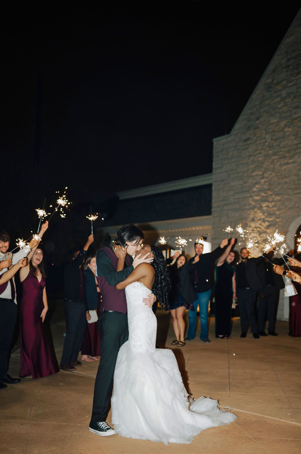 Wedding guests hold sparklers in a line at night while a bride and groom pose on the dance floor.