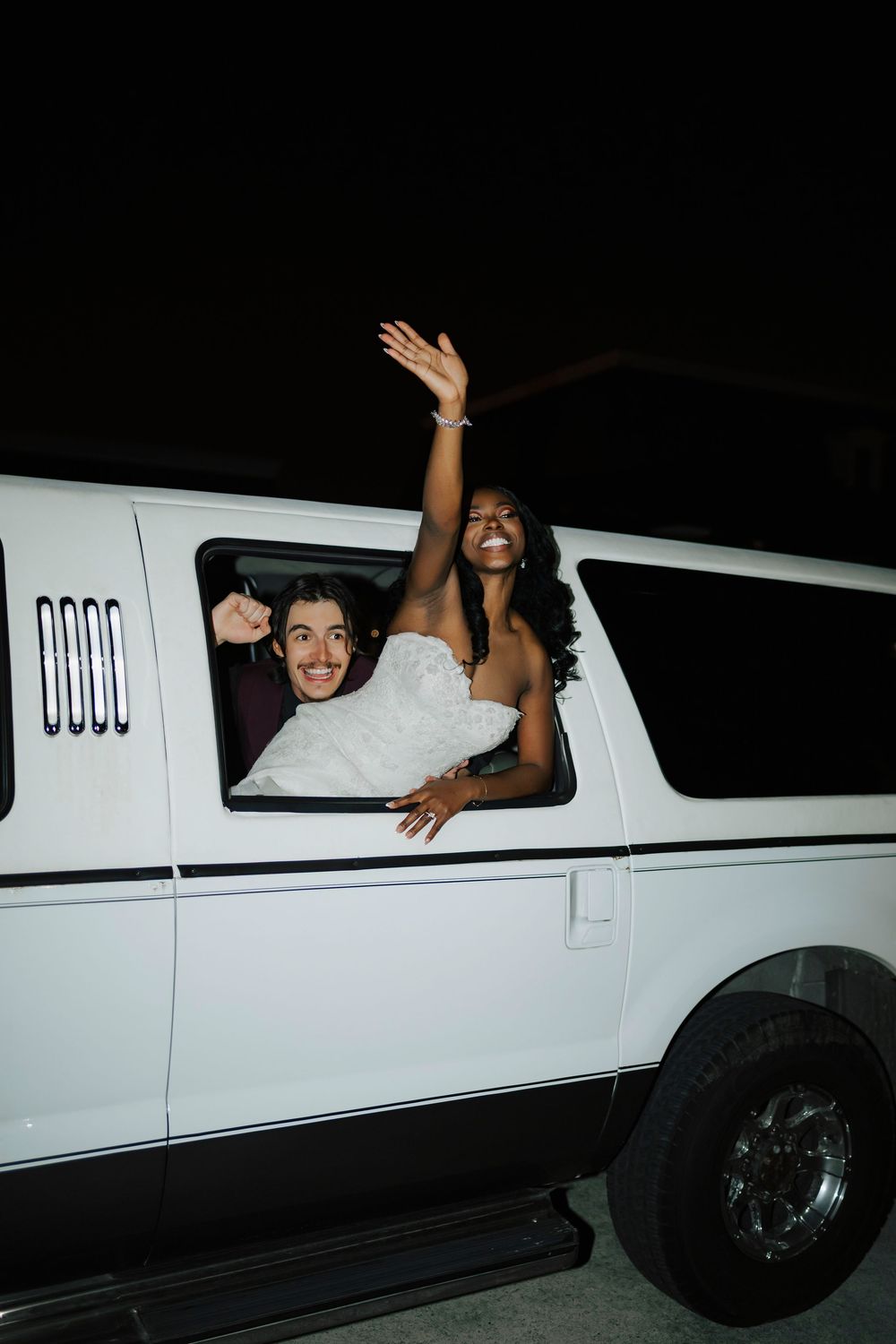 People waving and smiling from the window of a white limousine at night during a celebration.