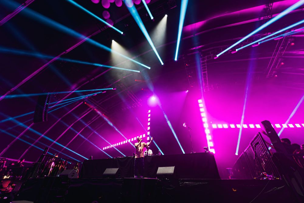 Concert venue illuminated with vibrant purple and blue laser lights creating dynamic patterns across a dark stage.