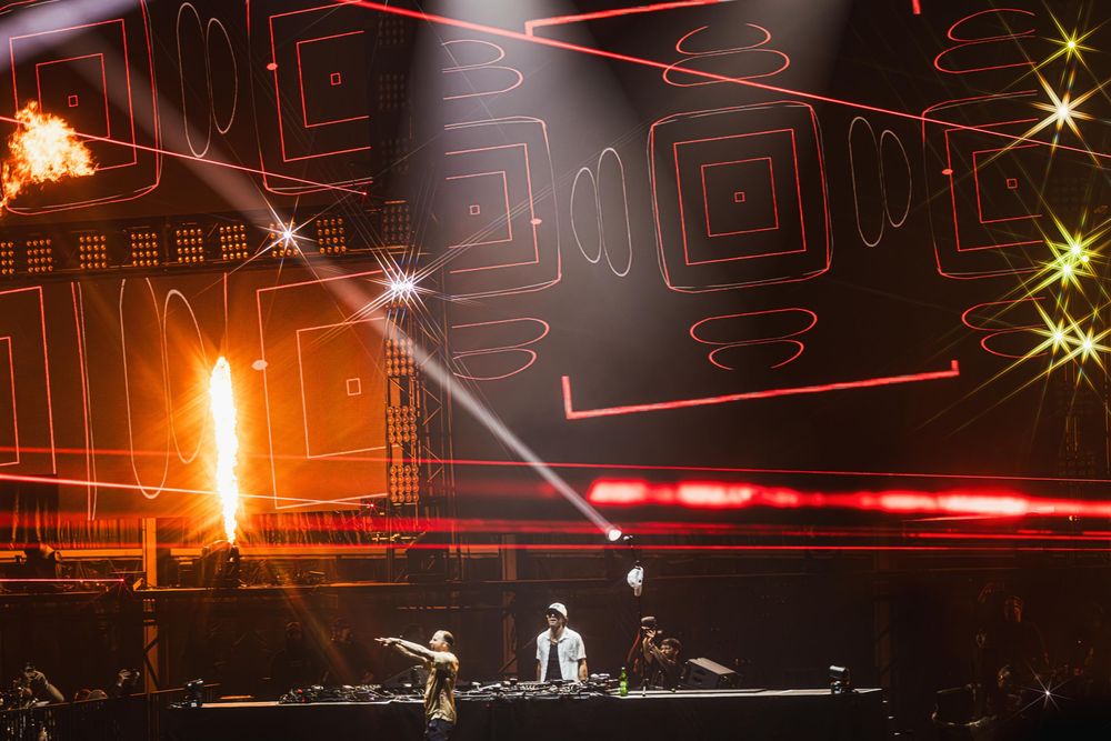 Colorful laser light show with geometric patterns illuminates a dark stage during a live performance.