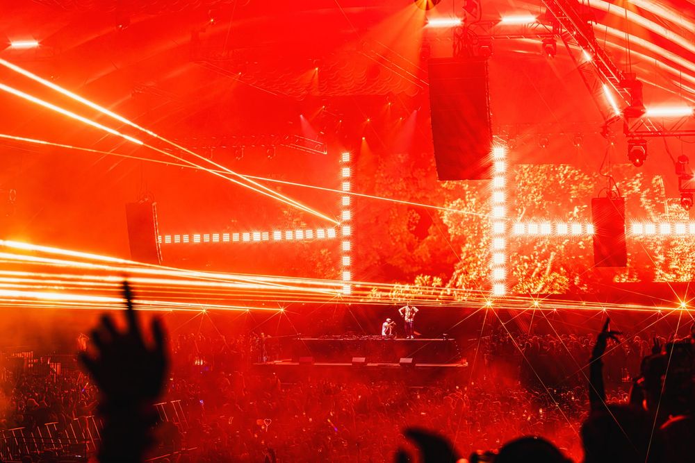 Concert crowd silhouettes under dramatic red lighting with bright laser beams creating an energetic atmosphere at live show.
