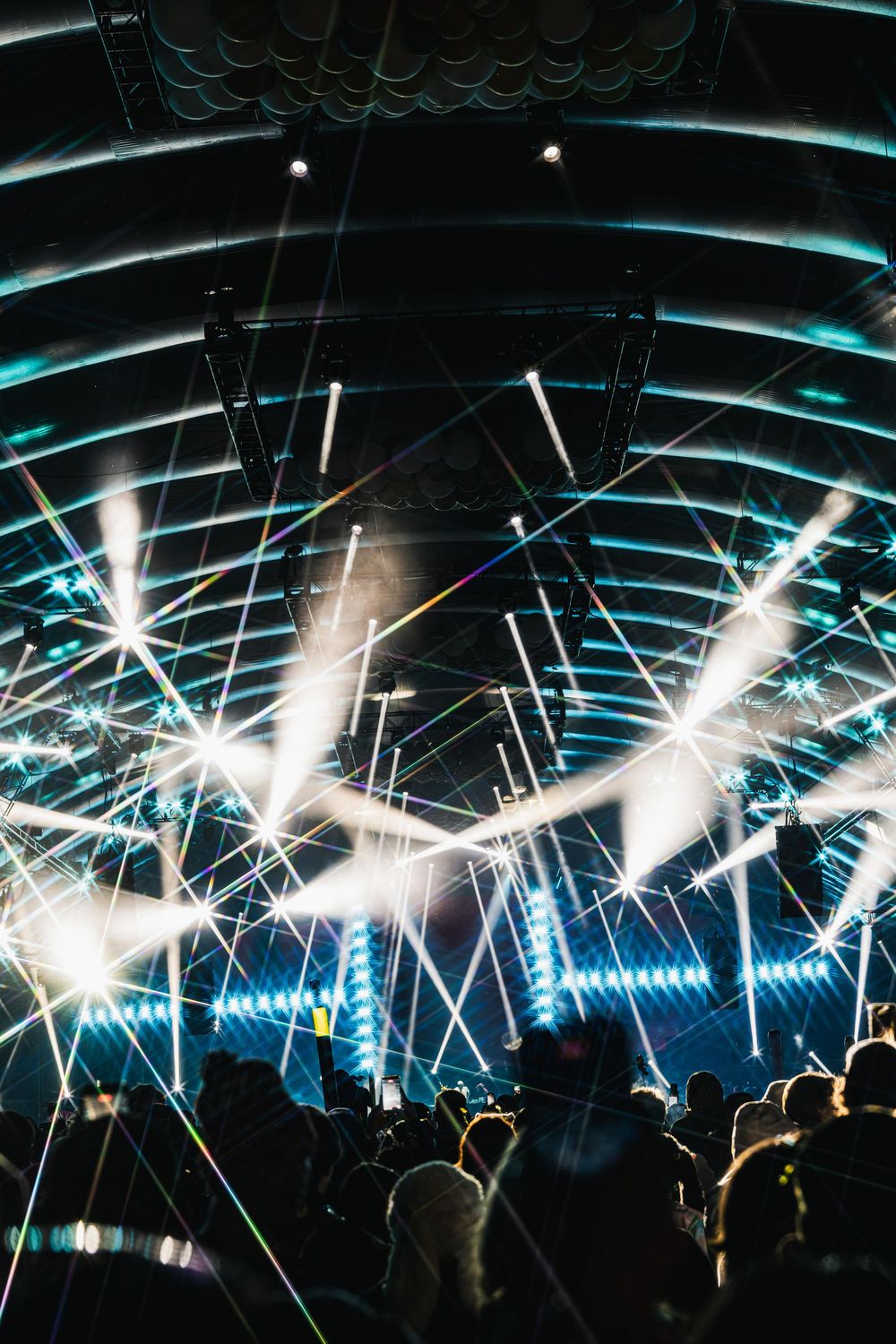 Electronic music festival stage illuminated with bright white strobe lights and turquoise ceiling beams over audience.