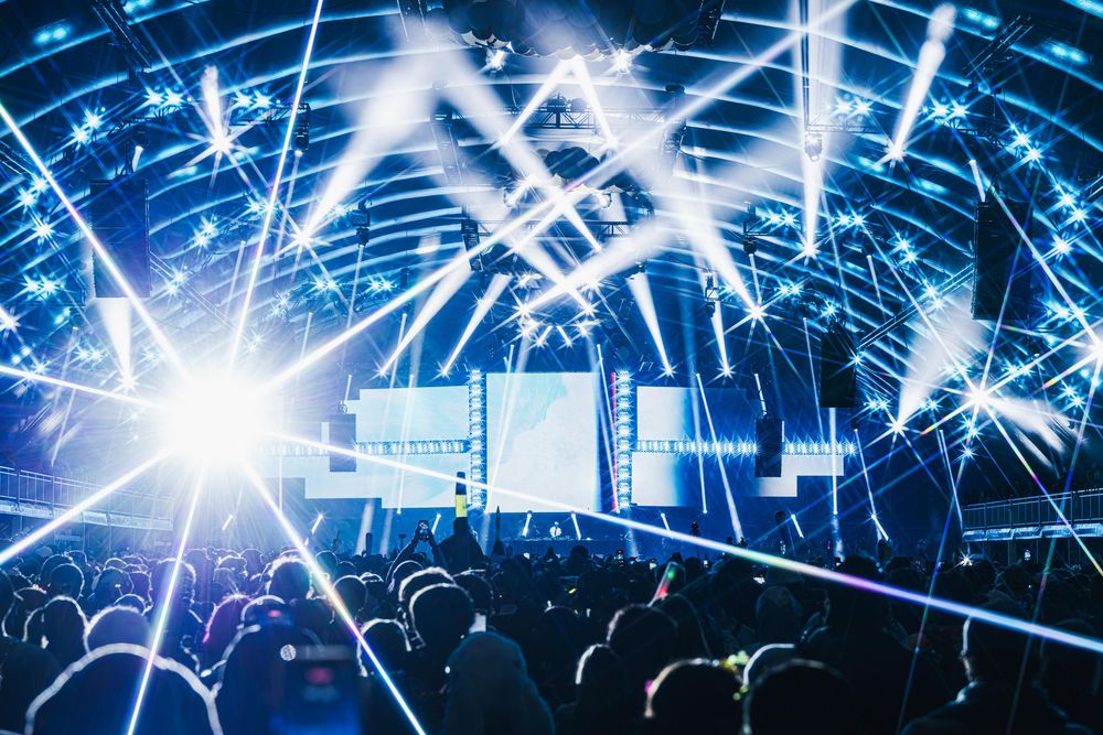 Concert crowd silhouettes against a backdrop of bright blue stage lights and laser beams creating an electric atmosphere.