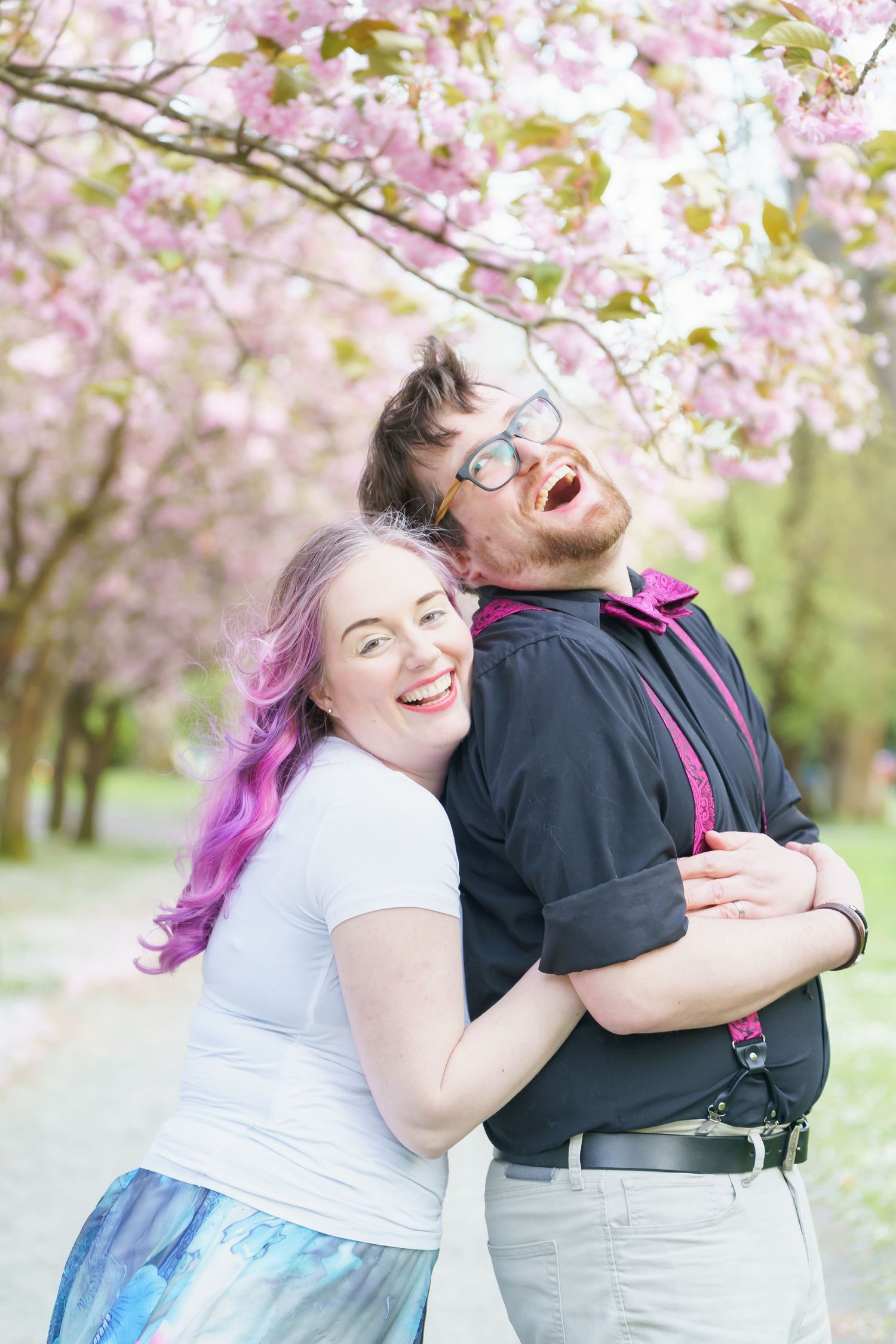 A couple laughs joyfully together under blooming pink cherry blossom trees in a spring garden setting.