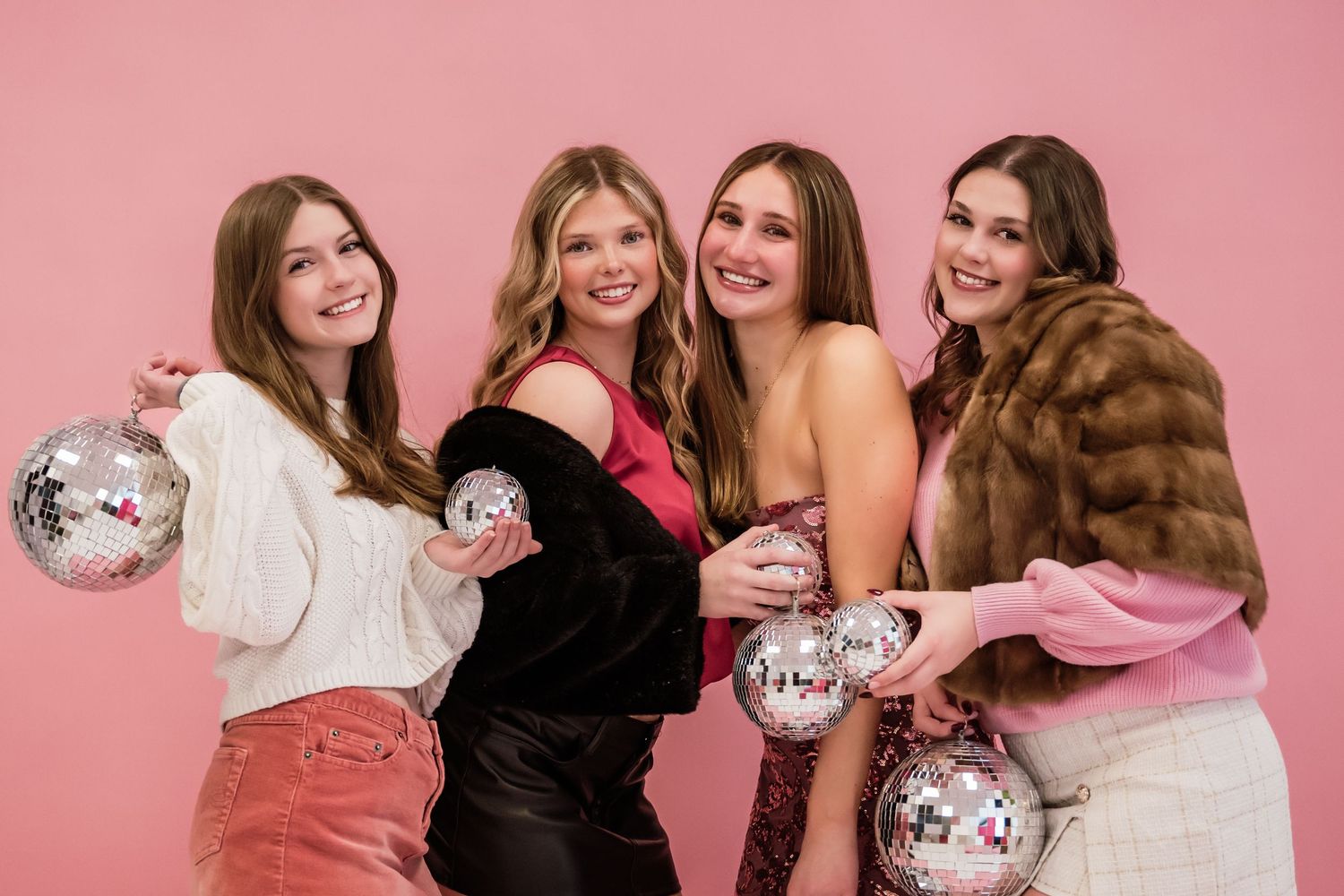 A group of friends pose with disco balls against a pink backdrop wearing stylish winter outfits and fur coats.
